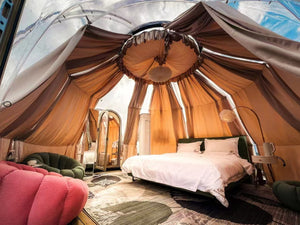 Interior view of a luxury glamping tent with elegant drapery and a cozy bed, complemented by plush seating and modern amenities, nestled within a transparent dome structure.