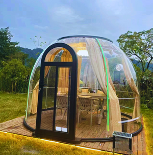 Transparent eco-friendly dome with wooden flooring and furnished interior, set in a lush green field with mountain views, ideal for sustainable travel and nature retreats.