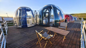 Panoramic view of multiple clear dome glamping pods on a wooden deck overlooking the ocean at sunset, equipped with outdoor chairs for a relaxing retreat at Earth Haven.